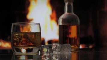 Glass and bottle of whiskey, scotch or bourbon stands on a table against background of a fireplace with a flame. Alcoholic drink and home comfort concept. video