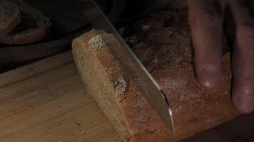 Cutting slice of homemade Crusty rye bread with a sharp knife on wooden board, closeup slow motion video