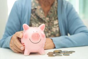 Asian elderly woman putting coin into pink piggy bank for saving money and insurance, poverty, financial problem in retirement. photo