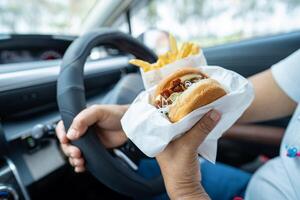 Asian lady holding hamburger and French fries to eat in car, dangerous and risk an accident. photo