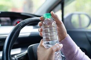 conductora asiática sosteniendo una botella para beber agua mientras conduce un automóvil. una botella de agua caliente de plástico provoca un incendio. foto