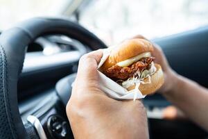 asiático dama participación hamburguesa a comer en auto, peligroso y riesgo un accidente. foto