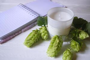 The opened notepad, pen, white candle, glasses and branches of hops as decoration on a white wooden table. Desktop still life with space for text. photo