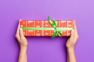 Flat lay of woman hands holding gift wrapped and decorated with bow on purple background with copy space. Christmas and holiday concept photo