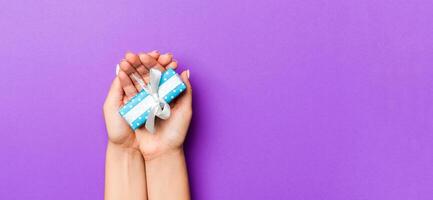 Flat lay of woman hands holding gift wrapped and decorated with bow on purple background with copy space. Christmas and holiday concept photo