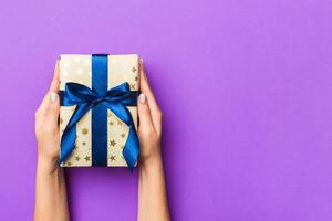 Flat lay of woman hands holding gift wrapped and decorated with bow on purple background with copy space. Christmas and holiday concept photo