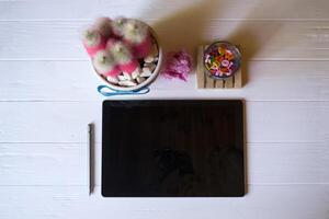 A tablet with stylus, pink cactus, crumpled sheet of paper and multicolored letters on a white wooden desktop. photo