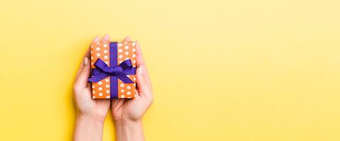 Woman arms holding gift box with colored ribbon on yellow table background, top view and copy space for you design photo