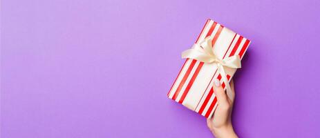 Flat lay of woman hands holding gift wrapped and decorated with bow on purple background with copy space. Christmas and holiday concept photo