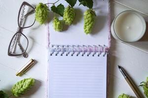 The opened notepad, pen, white candle, glasses and branches of hops as decoration on a white wooden table. Desktop still life with space for text. photo