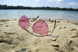 Pink glasses on the sand near blue water of lake. photo