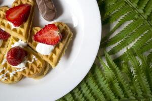 Waffles with strawberry on a plate, green leaves of fern on a table. Close up. Beautiful and tasty breakfast. photo