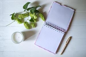 The opened notepad, pen, white candle, glasses and branches of hops as decoration on a white wooden table. Desktop still life with space for text. photo