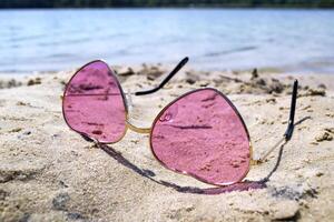 rosado lentes en el arena cerca azul agua de lago. foto