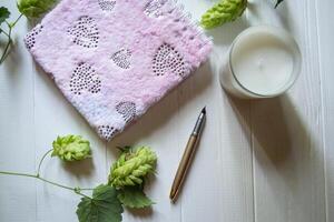 The closed pink notepad, pen, white candle and branches of hops as decoration on a white wooden table. Desktop still life. photo