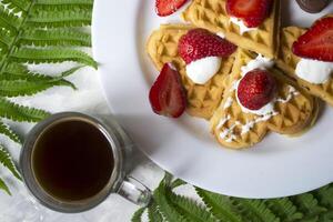 Waffles with strawberry, a cup of coffee and green leaves of fern on a table. Beautiful and tasty breakfast. photo