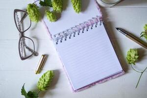 The opened notepad, pen, white candle, glasses and branches of hops as decoration on a white wooden table. Desktop still life with space for text. photo