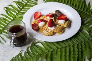 gofres con fresa, un taza de café y verde hojas de helecho en un mesa. hermosa y sabroso desayuno. foto