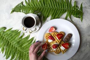 Waffles with strawberry, a cup of coffee and green leaves of fern on a table. Beautiful and tasty breakfast. photo