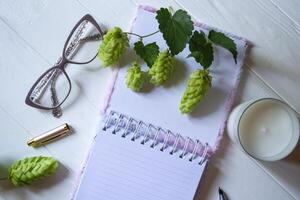 The opened notepad, pen, white candle, glasses and branches of hops as decoration on a white wooden table. Desktop still life with space for text. photo