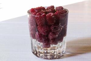 Ripe raspberries in a glass with water, on a white wooden background. photo