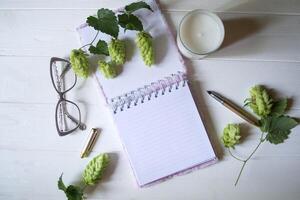 The opened notepad, pen, white candle, glasses and branches of hops as decoration on a white wooden table. Desktop still life with space for text. photo