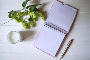 The opened notepad, pen, white candle, glasses and branches of hops as decoration on a white wooden table. Desktop still life with space for text. photo