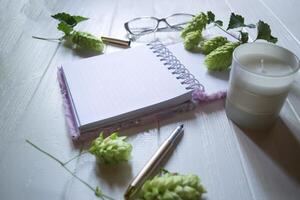 The opened notepad, pen, white candle, glasses and branches of hops as decoration on a white wooden table. Desktop still life with space for text. photo