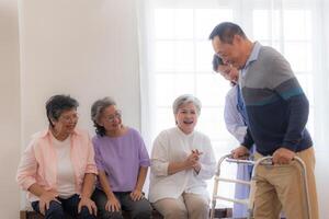 Asian young nurse support senior older man walk with walker. Beautiful nurse help and take care. elderly mature grandpa patient doing physical therapy with a group of senior friends in hospital. photo