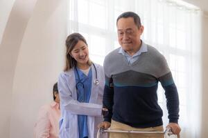 Asian young nurse support senior older man walk with walker. Beautiful nurse help and take care. elderly mature grandpa patient doing physical therapy with a group of senior friends in hospital. photo