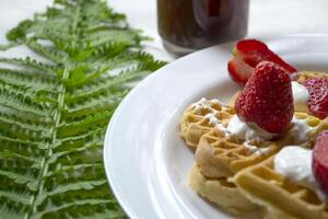 Waffles with strawberry, a cup of coffee and green leaves of fern on a table. Beautiful and tasty breakfast. photo