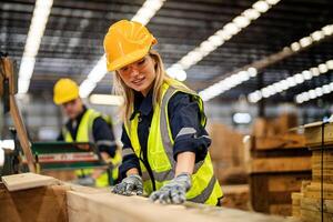 woman cleaning timber wood in dark warehouse industry. Team worker carpenter wearing safety uniform and hard hat working and checking the quality of wooden products at workshop manufacturing. photo