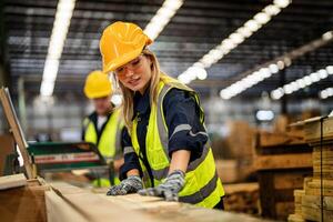 woman cleaning timber wood in dark warehouse industry. Team worker carpenter wearing safety uniform and hard hat working and checking the quality of wooden products at workshop manufacturing. photo