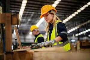 woman cleaning timber wood in dark warehouse industry. Team worker carpenter wearing safety uniform and hard hat working and checking the quality of wooden products at workshop manufacturing. photo