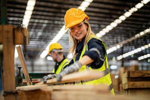 woman cleaning timber wood in dark warehouse industry. Team worker carpenter wearing safety uniform and hard hat working and checking the quality of wooden products at workshop manufacturing. photo