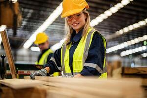woman cleaning timber wood in dark warehouse industry. Team worker carpenter wearing safety uniform and hard hat working and checking the quality of wooden products at workshop manufacturing. photo