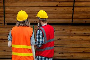 Team worker carpenter wearing safety uniform and hard hat working and checking the quality of wooden products at workshop manufacturing. man and woman workers wood in dark warehouse industry. photo