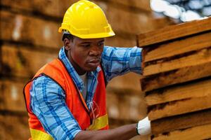 African worker carpenter wearing safety uniform and hard hat working and checking the quality of wooden products at workshop manufacturing. man and woman workers wood in dark warehouse industry. photo