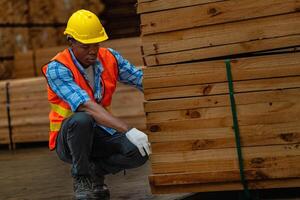 africano trabajador carpintero vistiendo la seguridad uniforme y difícil sombrero trabajando y comprobación el calidad de de madera productos a taller fabricación. hombre y mujer trabajadores madera en oscuro almacén industria. foto