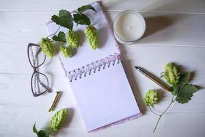 The opened notepad, pen, white candle, glasses and branches of hops as decoration on a white wooden table. Desktop still life with space for text. photo