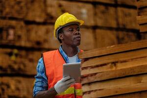 African worker carpenter wearing safety uniform and hard hat working and checking the quality of wooden products at workshop manufacturing. man and woman workers wood in dark warehouse industry. photo