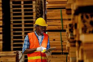 African worker carpenter wearing safety uniform and hard hat working and checking the quality of wooden products at workshop manufacturing. man and woman workers wood in dark warehouse industry. photo