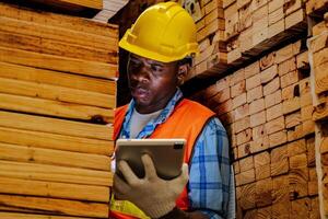 African worker carpenter wearing safety uniform and hard hat working and checking the quality of wooden products at workshop manufacturing. man and woman workers wood in dark warehouse industry. photo