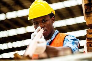 africano trabajador carpintero vistiendo la seguridad uniforme y difícil sombrero trabajando y comprobación el calidad de de madera productos a taller fabricación. hombre y mujer trabajadores madera en oscuro almacén industria. foto