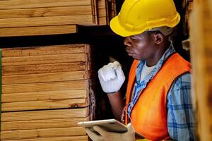 africano trabajador carpintero vistiendo la seguridad uniforme y difícil sombrero trabajando y comprobación el calidad de de madera productos a taller fabricación. hombre y mujer trabajadores madera en oscuro almacén industria. foto