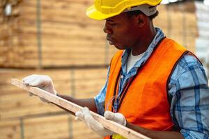 africano trabajador carpintero vistiendo la seguridad uniforme y difícil sombrero trabajando y comprobación el calidad de de madera productos a taller fabricación. hombre y mujer trabajadores madera en oscuro almacén industria. foto