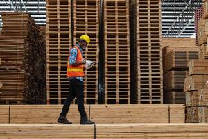 African workers man engineering walking and inspecting with working suite dress and hand glove in timber wood warehouse. Concept of smart industry worker operating. Wood factories produce wood palate. photo