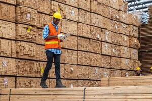 African workers man engineering walking and inspecting with working suite dress and hand glove in timber wood warehouse. Concept of smart industry worker operating. Wood factories produce wood palate. photo