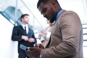 inteligente negro empresario acecho móvil teléfono. calvo adulto hombre vistiendo formal tener puesto. pensativo afroamericano empresario en un perfectamente a la medida traje en pie en escalera en oficina. foto