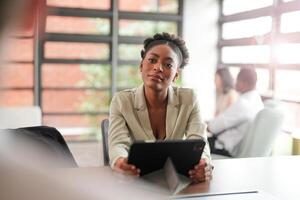 African black business woman using smartphone while working on laptop at office. Smiling mature african american businesswoman looking up while working on phone. Successful woman entrepreneur. photo